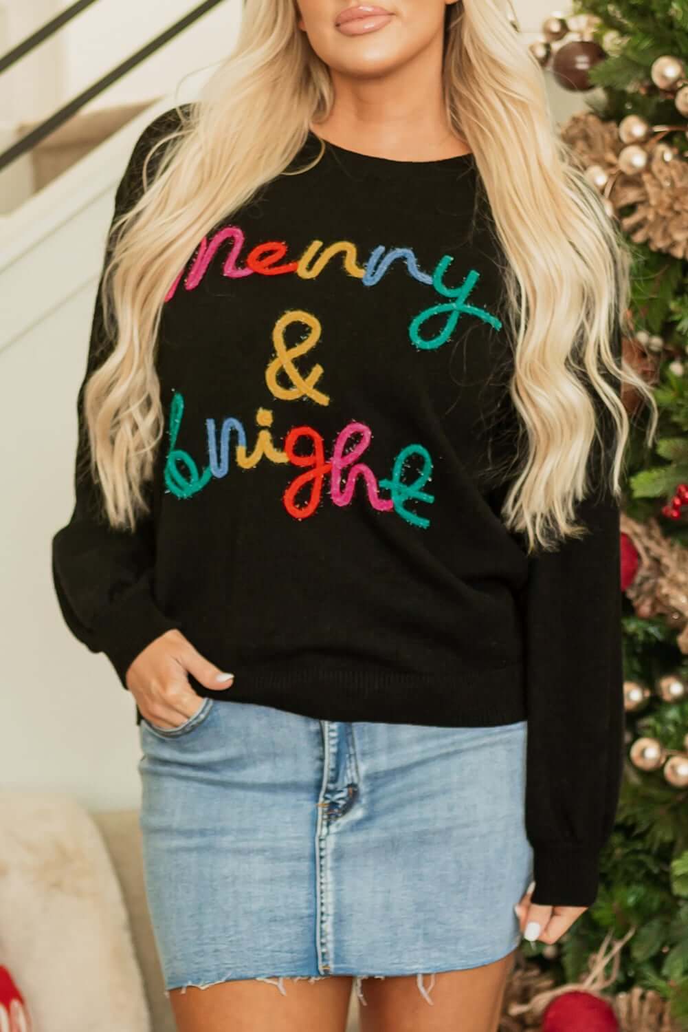 Model wearing MERRY & BRIGHT SWEATER by Vivian-Lu, a festive black sweater with colorful tinsel lettering.