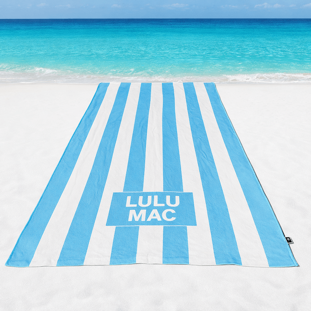 LULU MAC DARBY BEACH TOWEL (sky blue) on a sandy beach with turquoise ocean waves in the background.