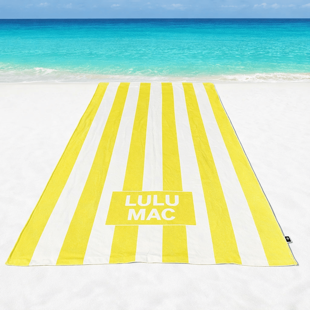 LULU MAC DARBY BEACH TOWEL (yellow) laid out on the beach with turquoise water in the background.