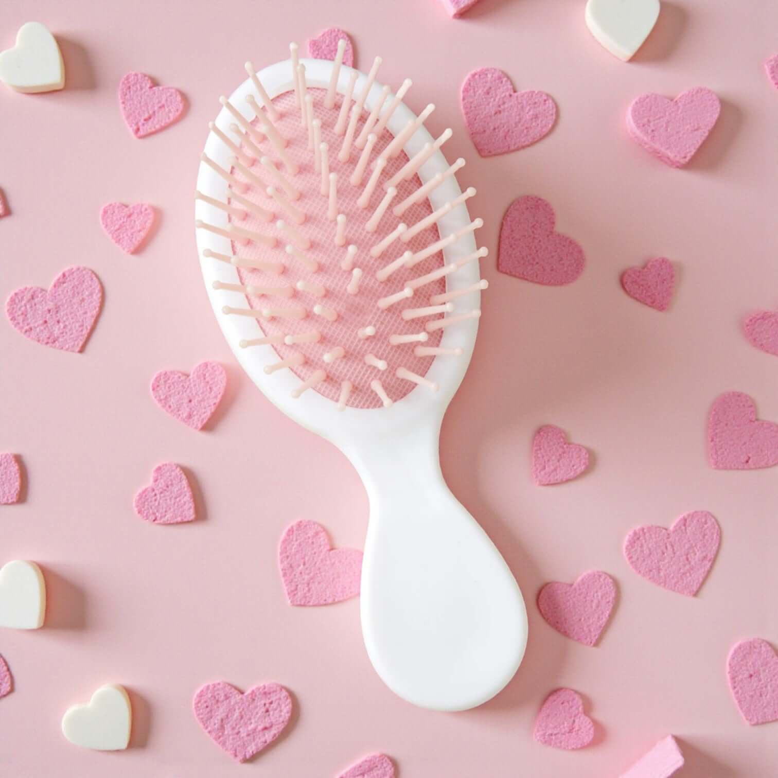 BOW BLISS MINI HAIRBRUSH on a pink background surrounded by heart-shaped candies, showcasing its cute design.
