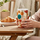 Person holding a colorful SWIG FALL HARVEST TRAVEL MUG 18 oz. next to a croissant on a wooden table.