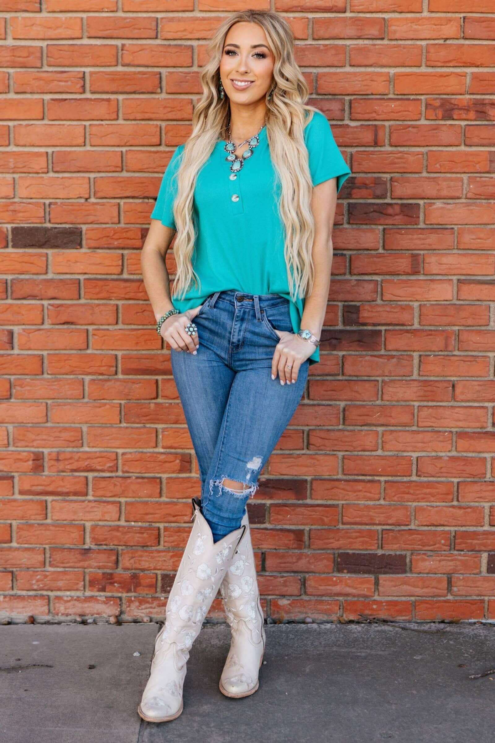 Woman wearing aqua Everyday Energy Top by Andree, styled with jeans and cowboy boots against a brick wall.