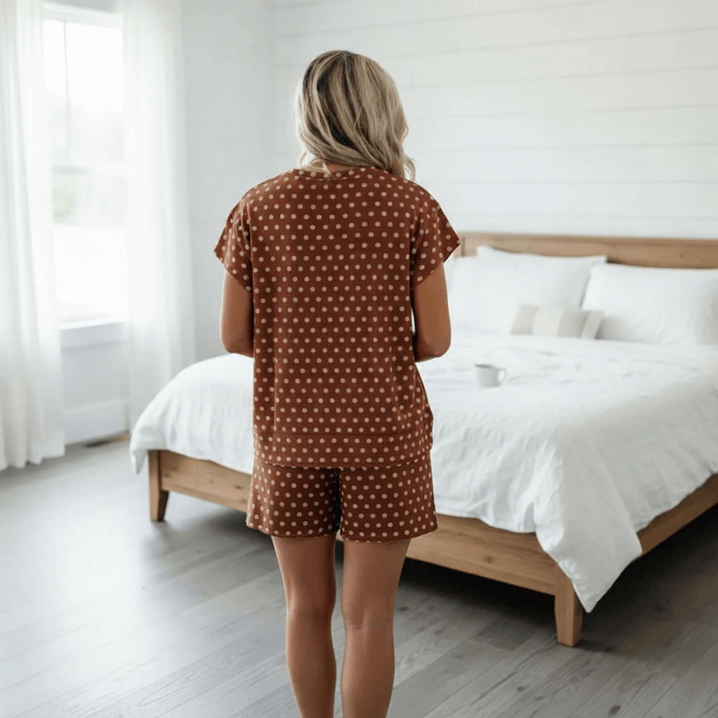 Woman wearing ON THE DOT SHORTS SET in mocha polka dots, relaxing in a bright bedroom.