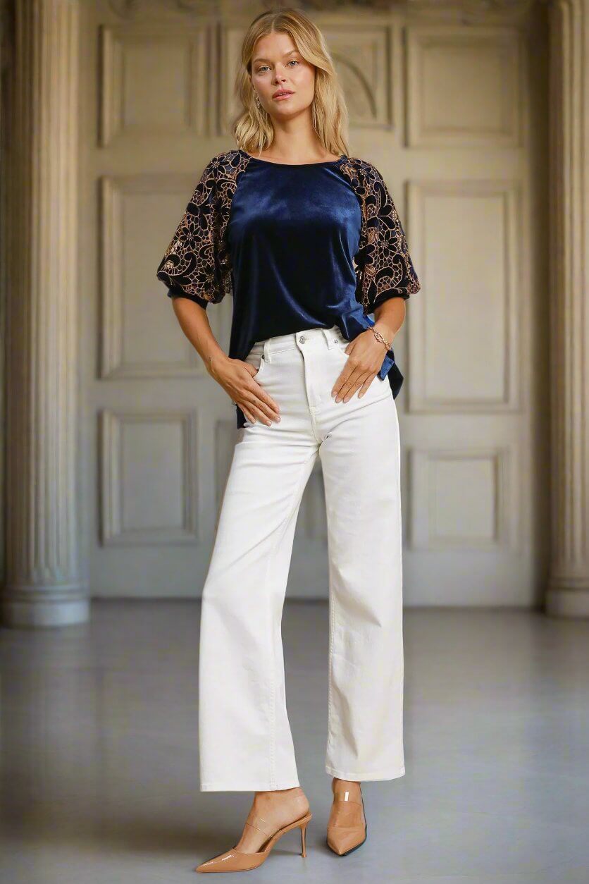 Elegant woman in navy blue velvet top with floral lace sleeves, paired with white wide-leg jeans and heels.