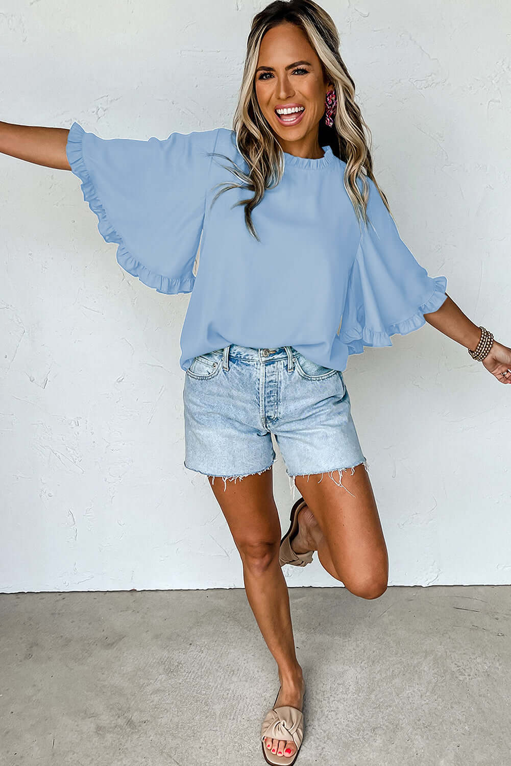 Model wearing a light blue Frill Seeker Top with ruffle trim, paired with denim shorts and sandals.
