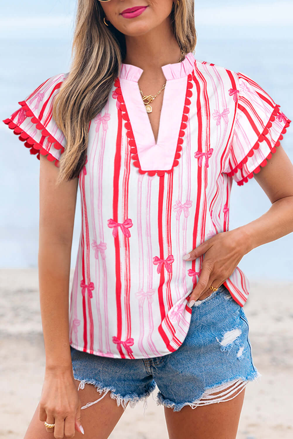 Model wearing the Ribbon Reign Top with bow print and ric-rac trim, paired with denim shorts, against a beach background.