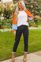 Model wearing the CITRUS CRUSH TOP with orange and blue color block sleeves, paired with dark jeans in a garden setting.