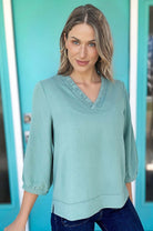 Model wearing GRACEFUL GLOW TOP (jade sea) with frilled V-neckline and bracelet sleeves, against a turquoise background.