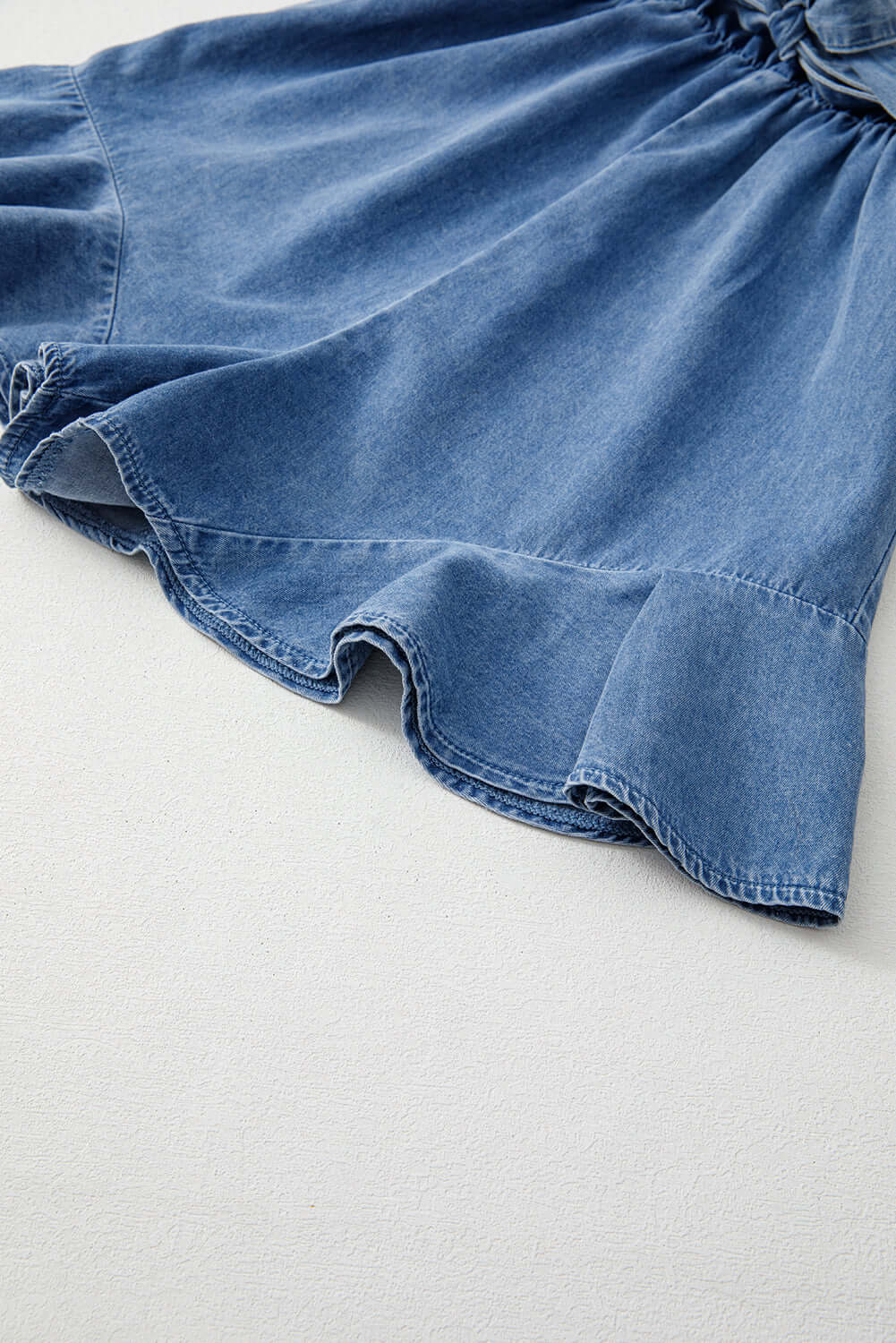 Lightweight medium blue denim romper featuring a feminine ruffled hem and breathable fabric for warm weather comfort.
