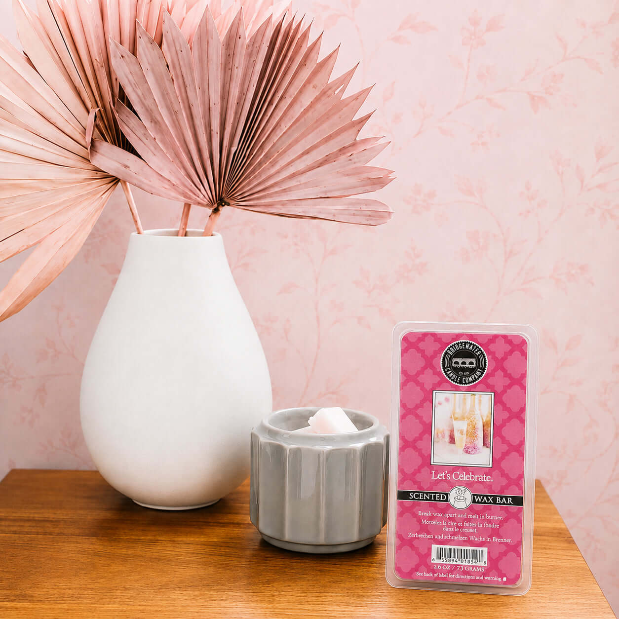 LETS CELEBRATE WAX MELTS displayed next to a vase with decorative leaves on a wooden table.