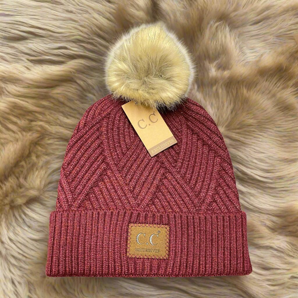 Warm angle beanie in burgundy with faux fur pom pom and unique diagonal pattern on a cozy background.