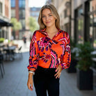 Model wearing the LAVA LUXE BLOUSE - JODIFL in vibrant red, orange, and purple tones, showcasing its silky fabric in an outdoor setting.