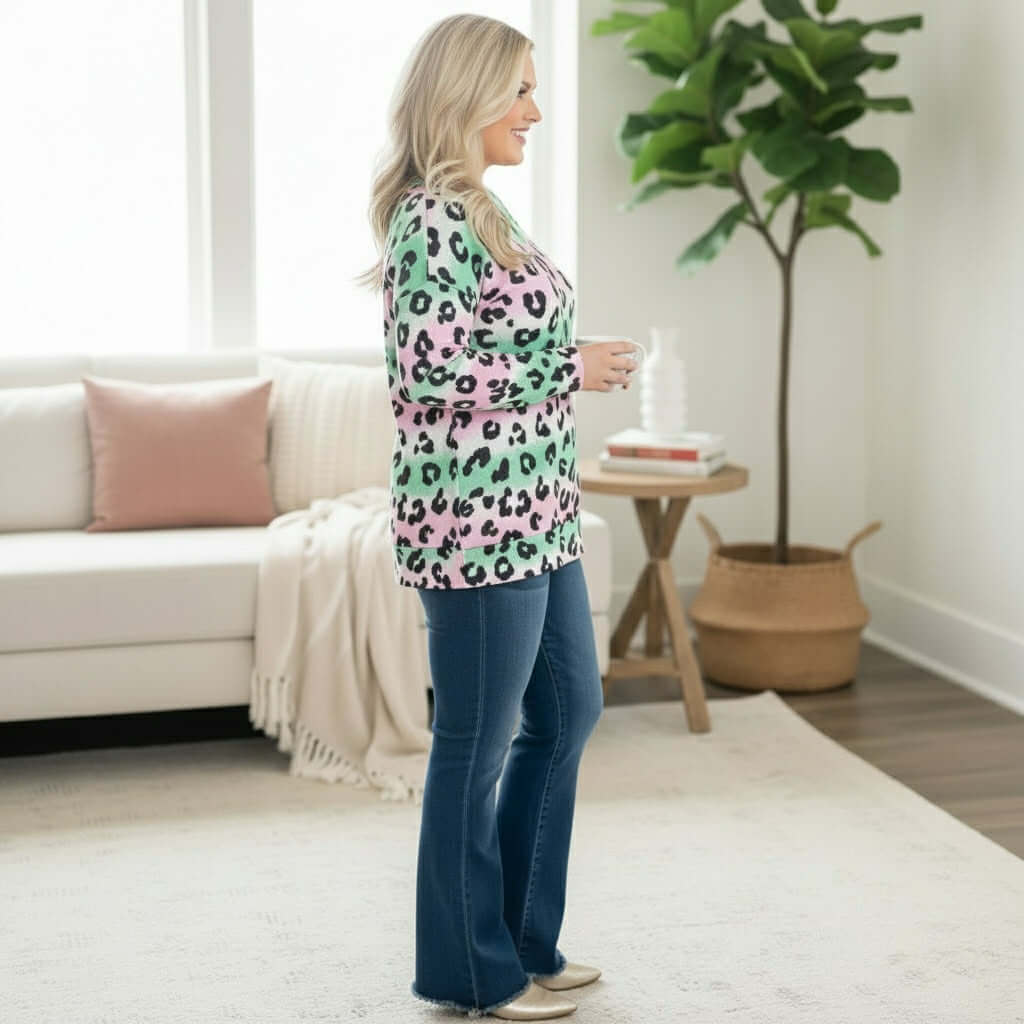 Woman wearing PRETTY PROWL TOP - Zenana in pastel leopard print, styled with flared jeans in a cozy living room setting.
