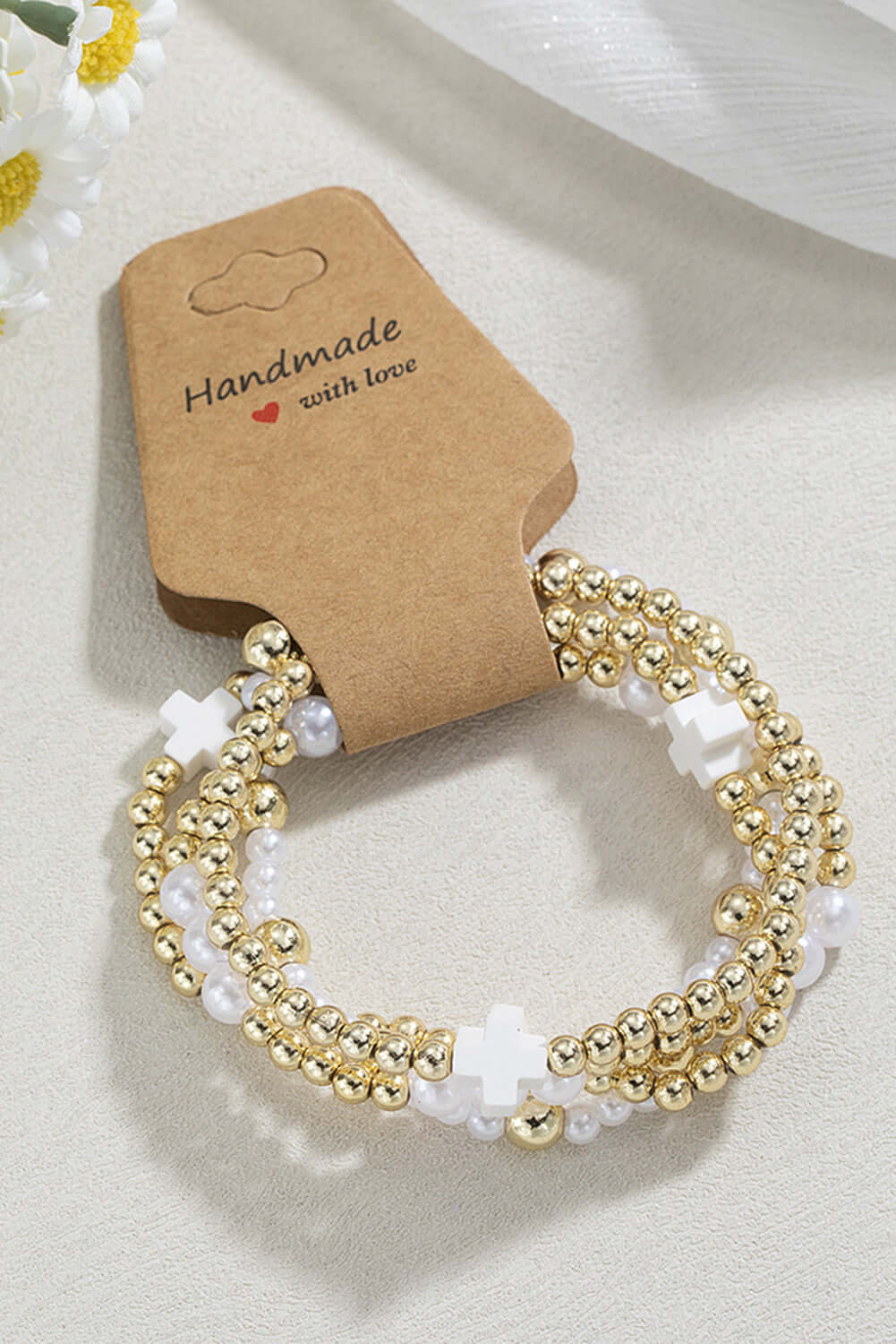 Handmade Timeless Faith bracelet stack with gold beads and white crosses displayed on a cream surface.