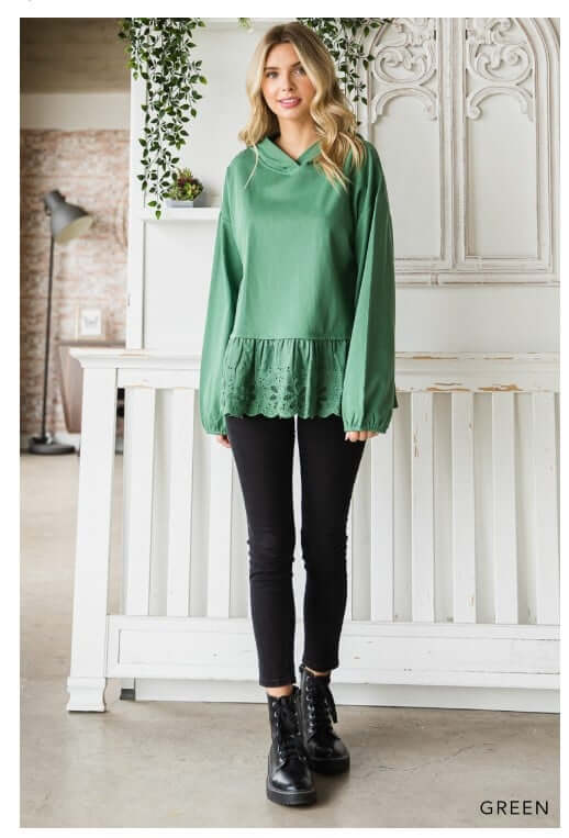 Woman in green ADVENTURE AWAITS hoodie top by Cotton Bleu with eyelet ruffle, styled with black pants and boots.