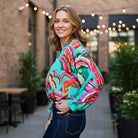 A smiling model wearing the LAGUNA LUXE BLOUSE - JODIFL in vibrant coastal colors in an outdoor setting.