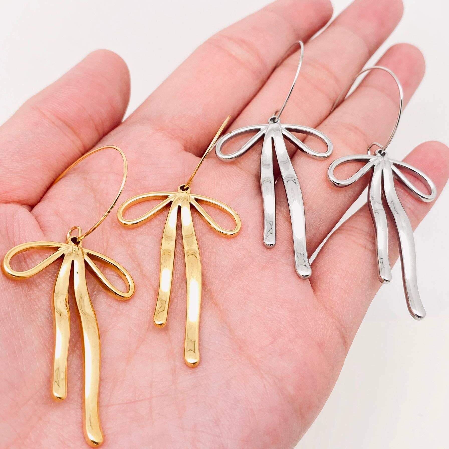 REFIEND RIBBON EARRINGS (2 colors) displayed in a hand, featuring gold and silver bow-inspired designs.