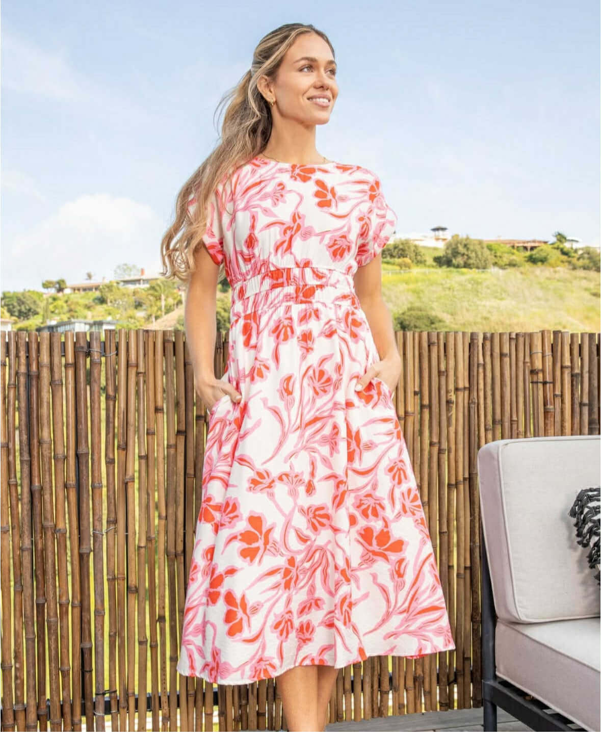 Woman modeling vibrant floral midi dress with pockets against a scenic outdoor backdrop.