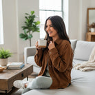Smiling woman wearing a Cinnamon Ridge Jacket, holding a coffee mug, sitting on a couch in a cozy living room.