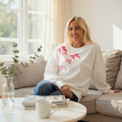Model wearing the DARLING DETAILS TOP, a white sequin bowknot oversized sweatshirt, sitting on a cozy couch.