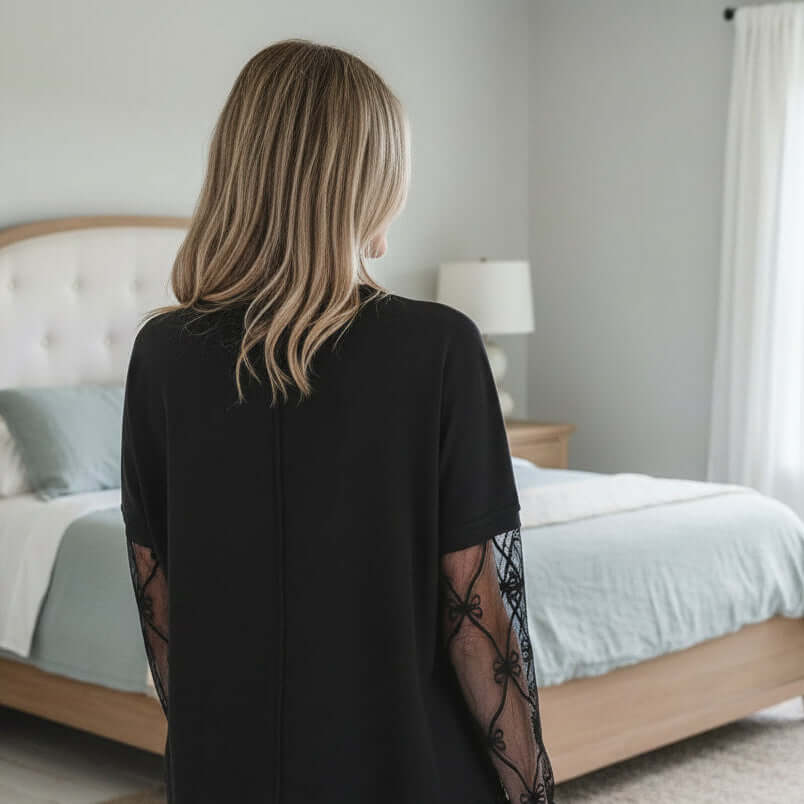 Woman wearing the SIMPLY ENCHANTED TOP, showcasing its sheer lace mesh sleeves in a cozy bedroom setting.