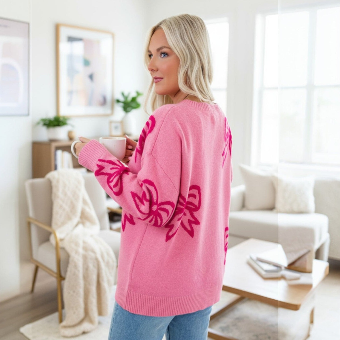 Woman wearing a RIBBON ROMANCE SWEATER with a pink bow pattern, enjoying coffee in a cozy living room.