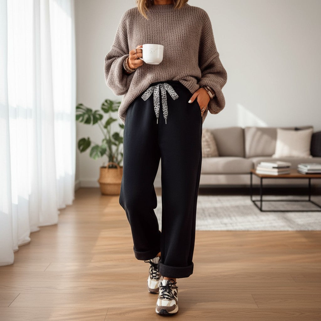 Woman in cozy sweater and COMFORT CULTURE PANTS (BLACK) with leopard drawstring, holding a coffee cup in stylish living room.