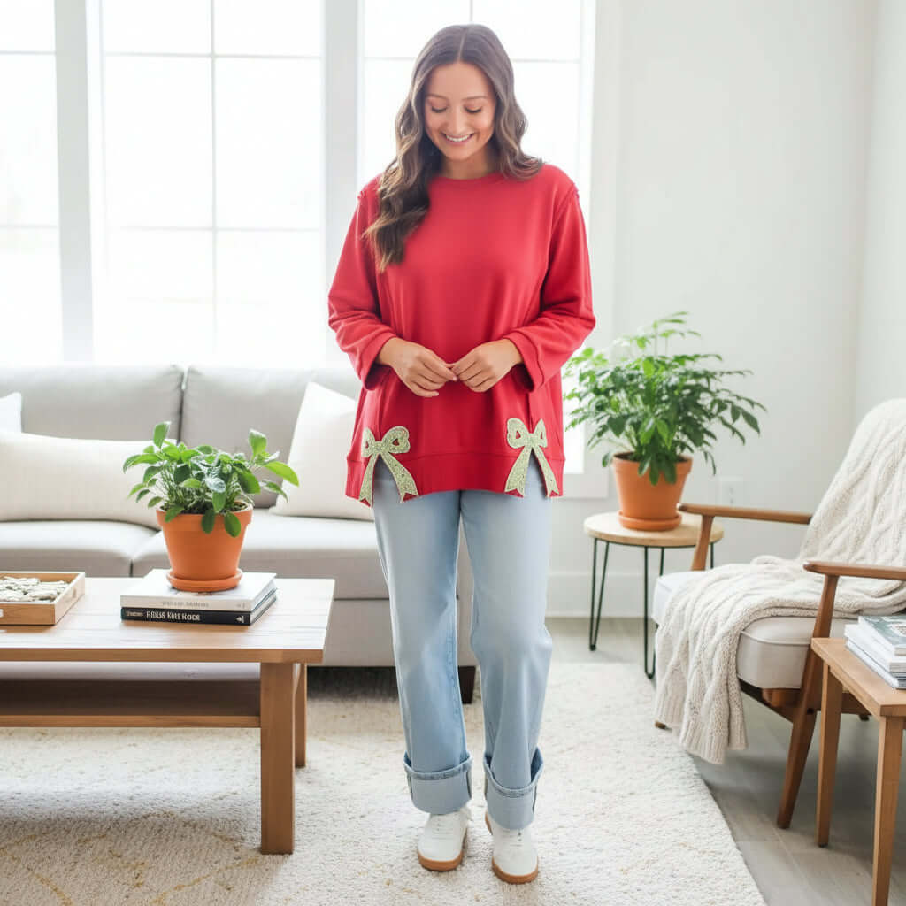 Model wearing SWEETHEART BOW TOP (red) with bow details, paired with relaxed jeans in a cozy living room setting.