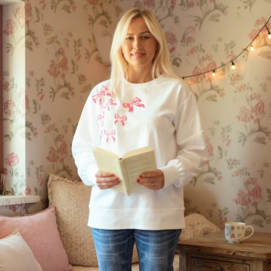 Model wearing the DARLING DETAILS TOP, a white oversized sweatshirt with sequin bows, reading a book in a cozy setting.