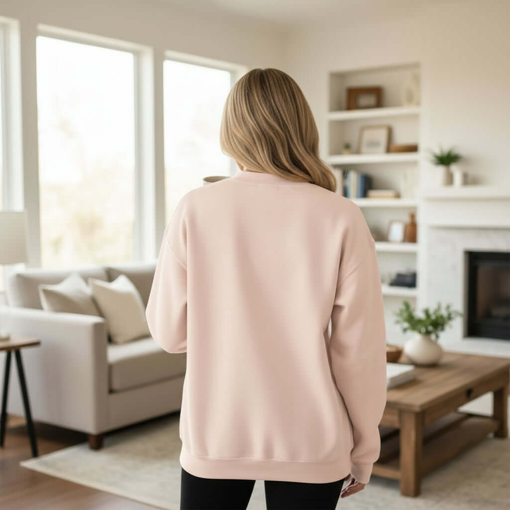 Back view of a woman wearing the LOVE LIKE JESUS PULLOVER in light pink, cozy setting with modern decor.