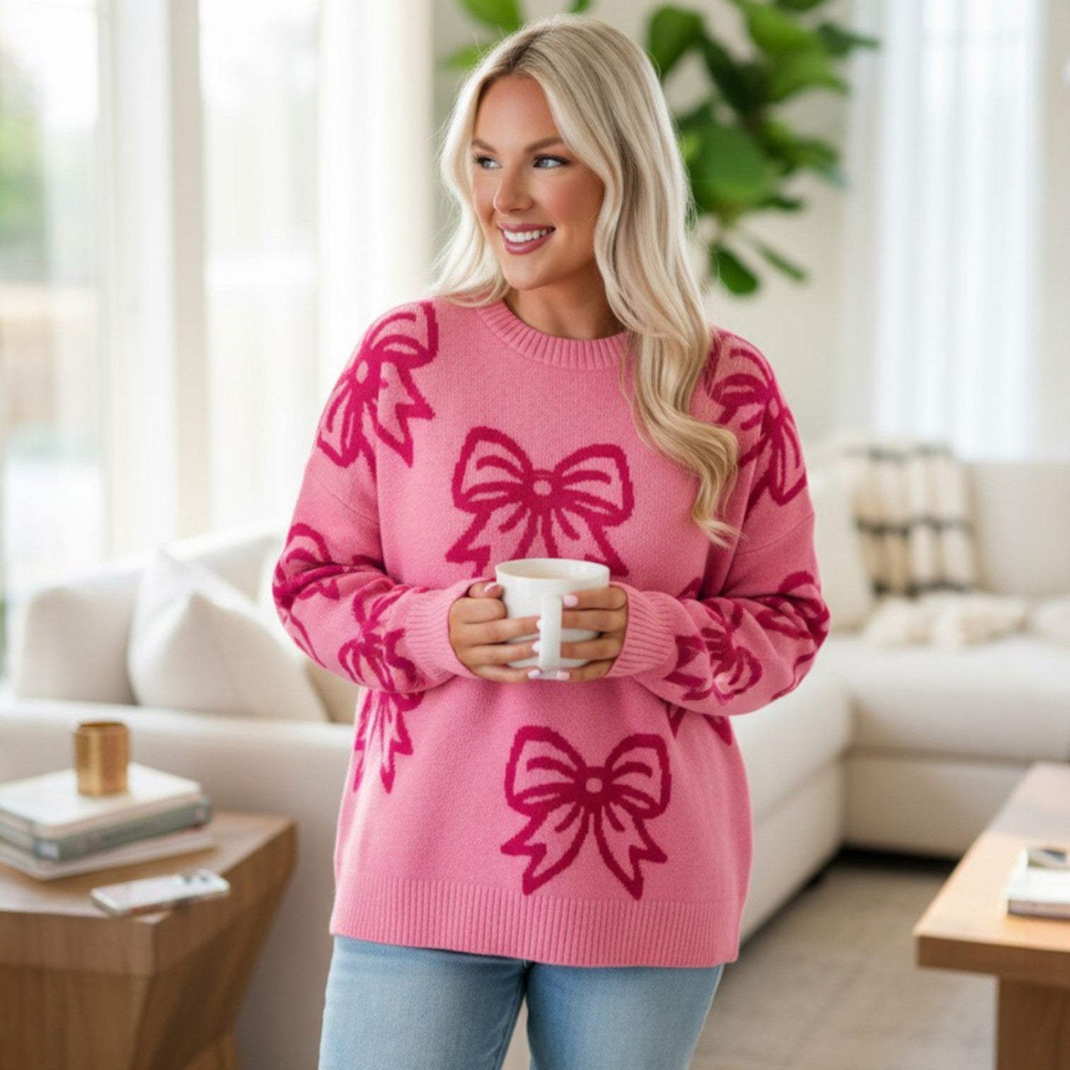Woman wearing RIBBON ROMANCE SWEATER with pink bow pattern while holding a coffee mug in a cozy living room.