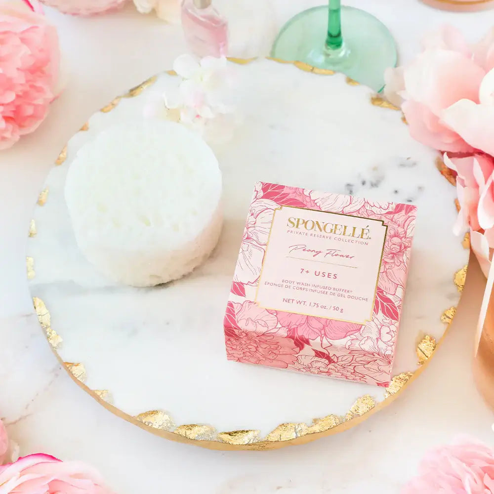 Private Reserve Peony Spongette on a marble tray surrounded by pink peonies, showcasing luxury body care.
