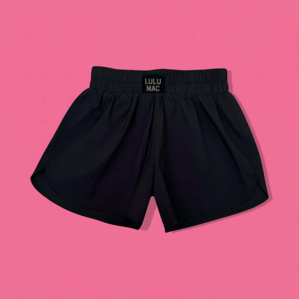 LULU MAC ACTIVE SHORTS (black) laid flat on a pink background, showcasing their lightweight and sporty design.