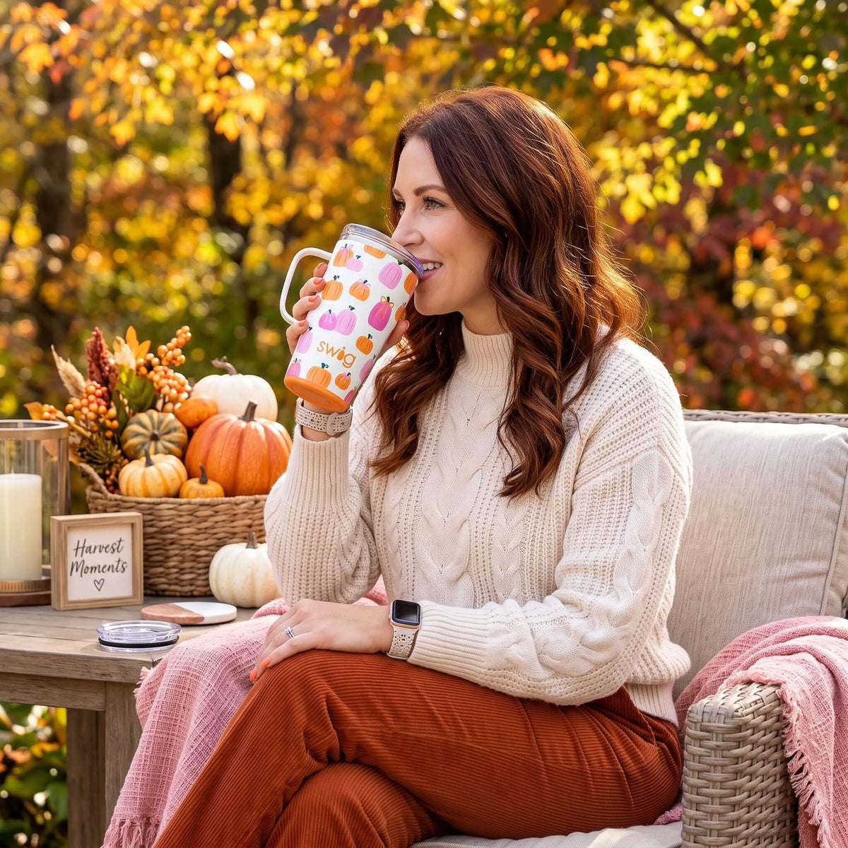 Woman enjoying a drink from PUMPKINS & BOWS TRAVEL MUG in a cozy fall setting with pumpkins and warm colors.