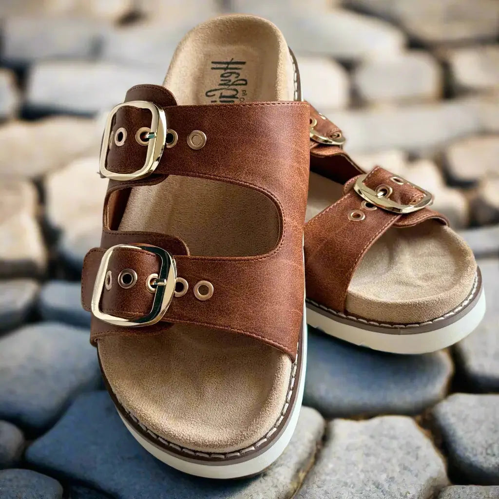 Feelin' Saucy sandals by Corkys with adjustable straps and shiny gold buckles on a cobblestone background.