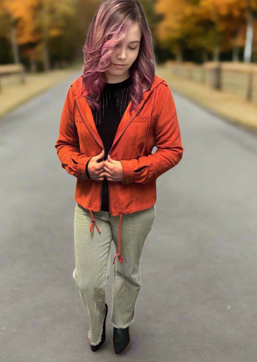 Woman wearing an Autumn Sunrise Jacket in ginger, standing on a fall path with colorful foliage.