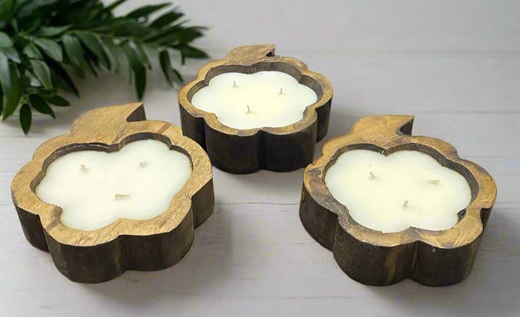 Three rustic pumpkin-shaped hand-poured soy candles with cotton wicks on a wooden surface, surrounded by greenery.