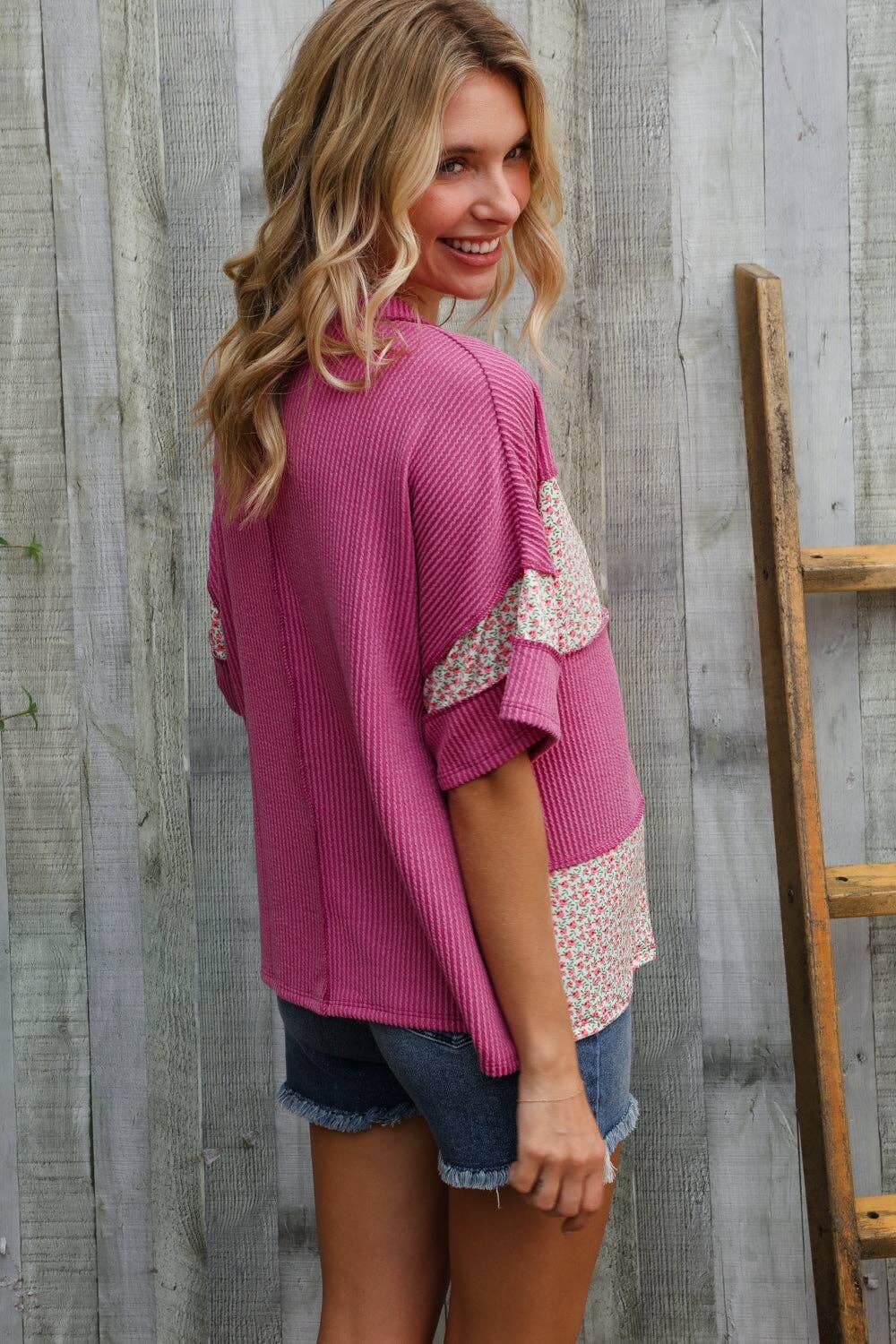Woman in fuchsia floral dolman pullover with color block design, styled with denim shorts, outdoors.