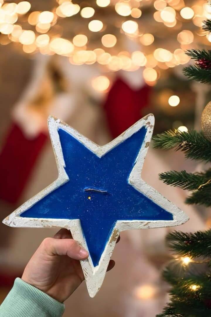 Hand holding a BLUE STAR CANDLE with rustic design, surrounded by festive lights and decorations, showcasing farmhouse charm.