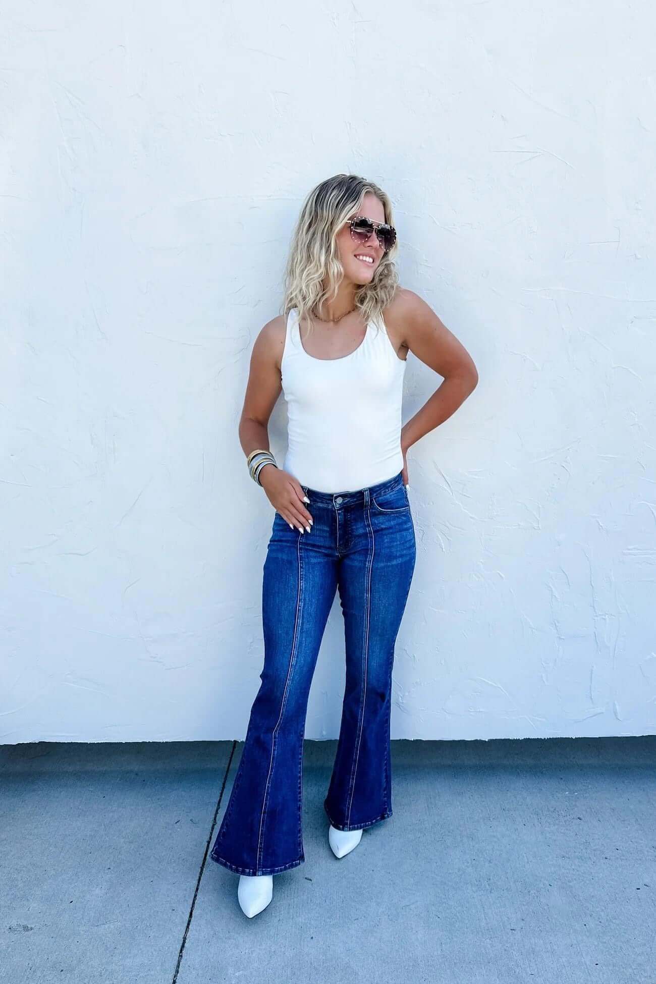 Model wearing mid-rise flare jeans with tummy control, styled with a white tank top and white heels.