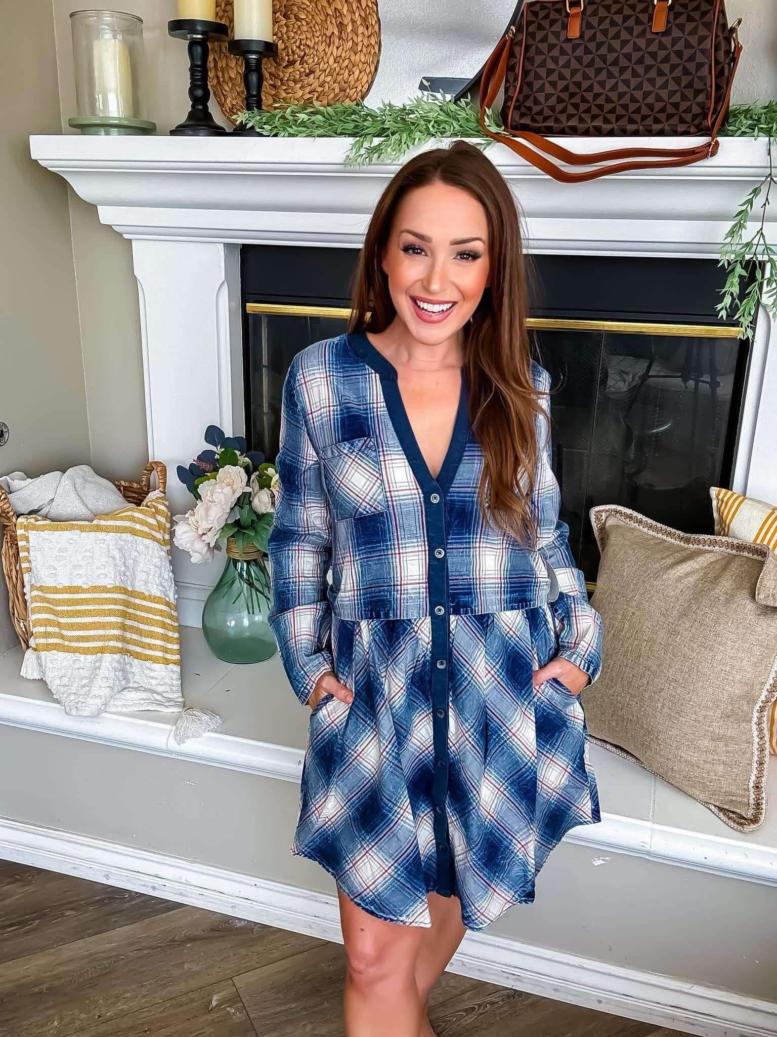 Woman wearing a blue plaid dress with buttons, standing by a fireplace in a cozy room.