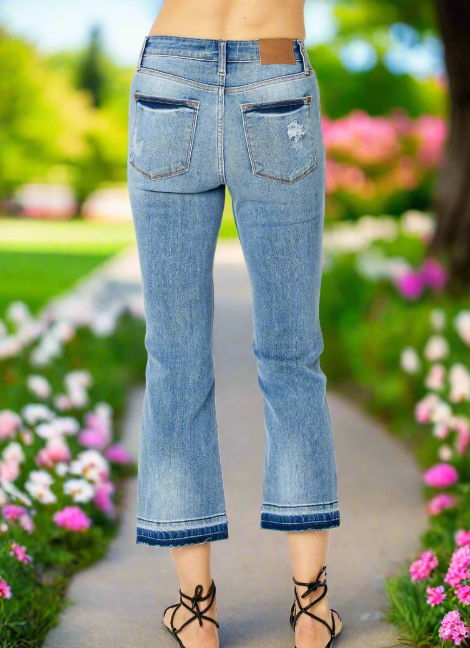 Model showcasing the back view of JUDY BLUE Amelia cropped bootcut jeans in light blue, surrounded by colorful flowers.