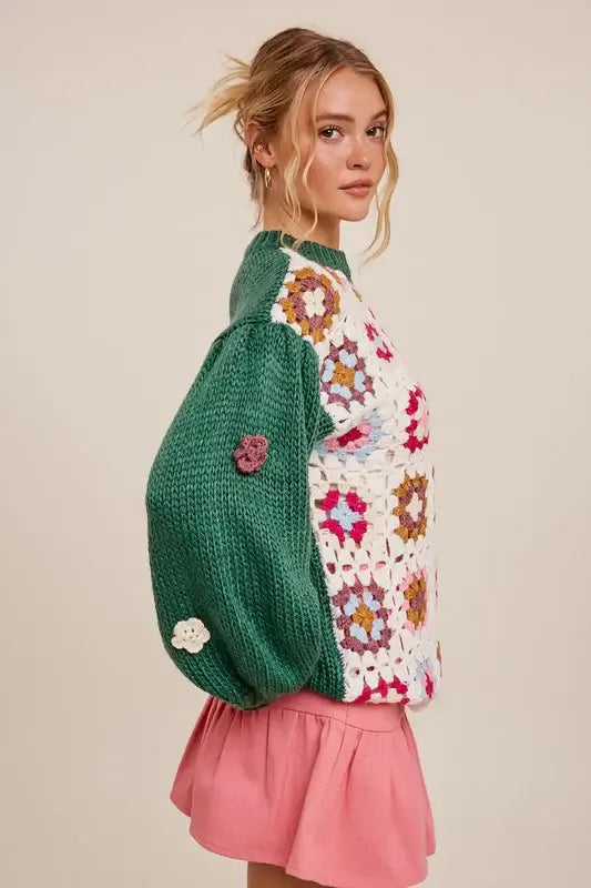 Boho floral patchwork sweater with 3D crocheted flowers, dark green sleeves, and a pink skirt modeled by a woman.