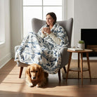 Cozy woman enjoying a warm drink in the SNOWY MOUNTAIN PLUSH THROW BLANKET while sitting in an elegant chair with a golden retriever.