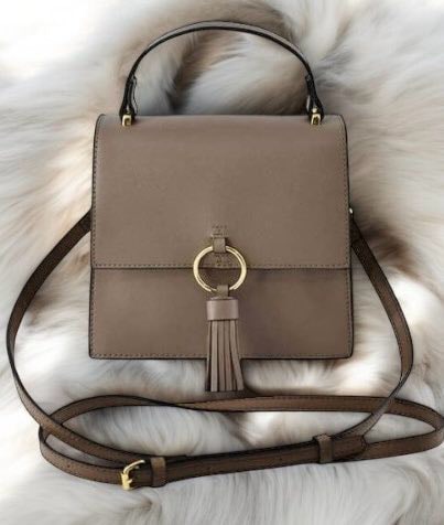 Taupe faux vegan leather crossbody bag with ring and tassel design, gold-tone hardware, detachable strap on soft fur background.