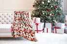 Cozy Christmas Cheer Plush Throw Blanket by Kathy Ireland draped over sofa, with festive design, near decorated tree and wrapped gifts.