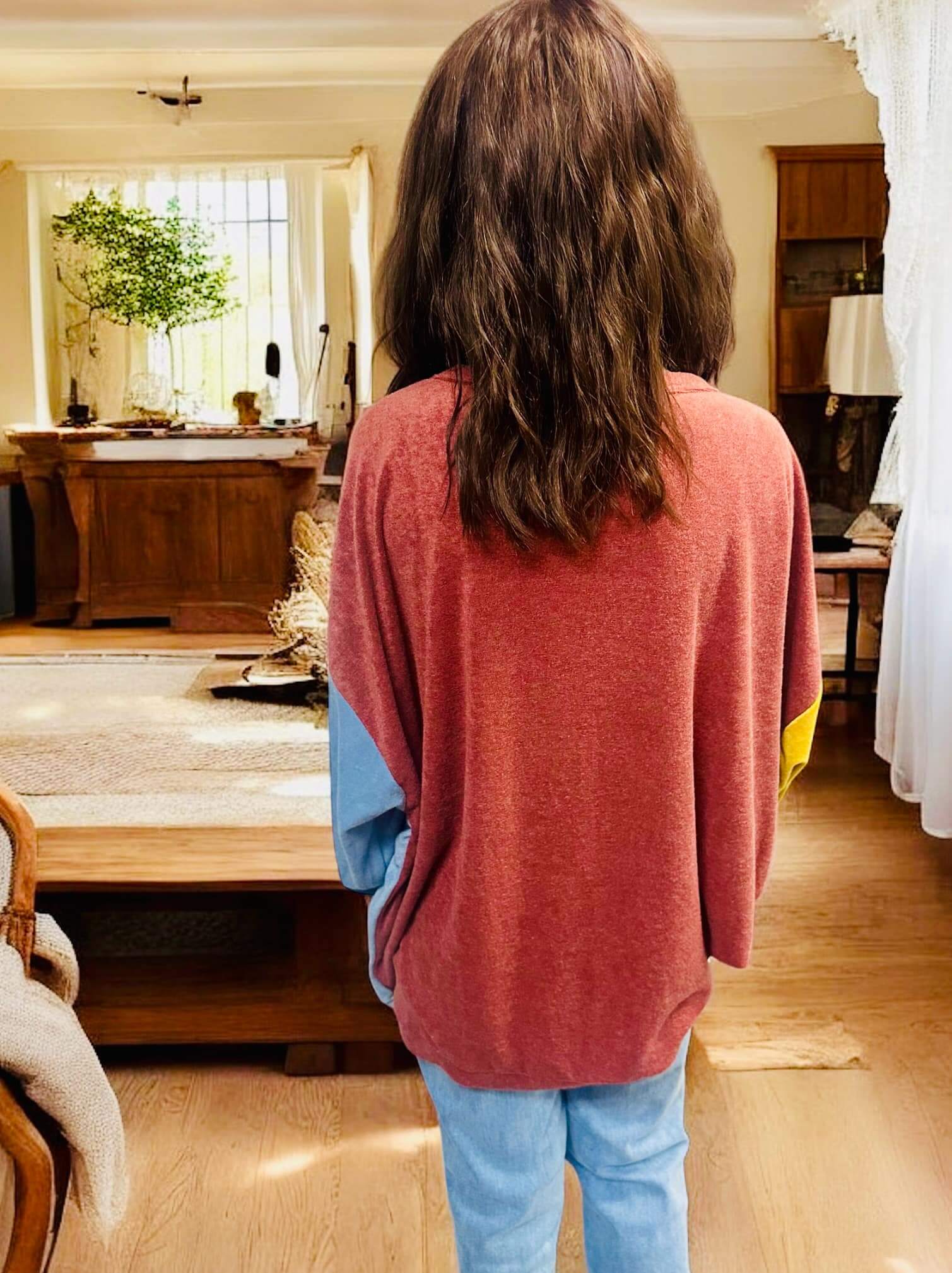 Woman wearing TRAVELING LIGHT top by BiBi in burgundy, gold, and blue color block design, standing in a cozy living room.