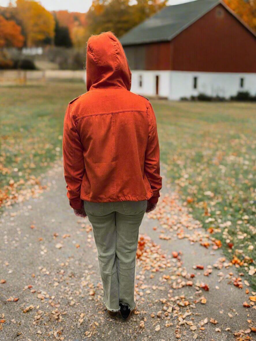 Person wearing Autumn Sunrise Jacket in ginger, walking on a leaf-covered path surrounded by fall colors, near a barn.