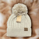 Tan cable knit beanie with pom pom and CC suede patch on faux fur background.