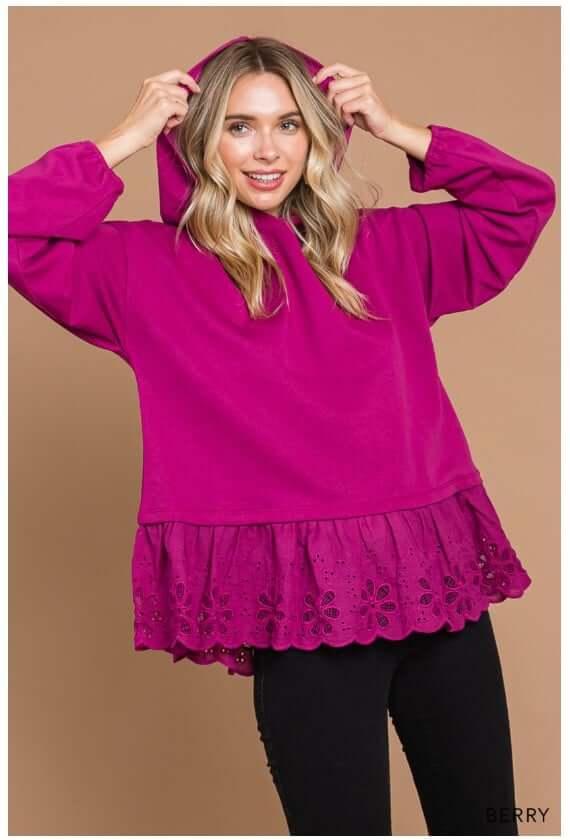 Woman wearing ADVENTURE AWAITS Berry Hoodie Top by Cotton Bleu with eyelet ruffle detail and cozy oversized design.